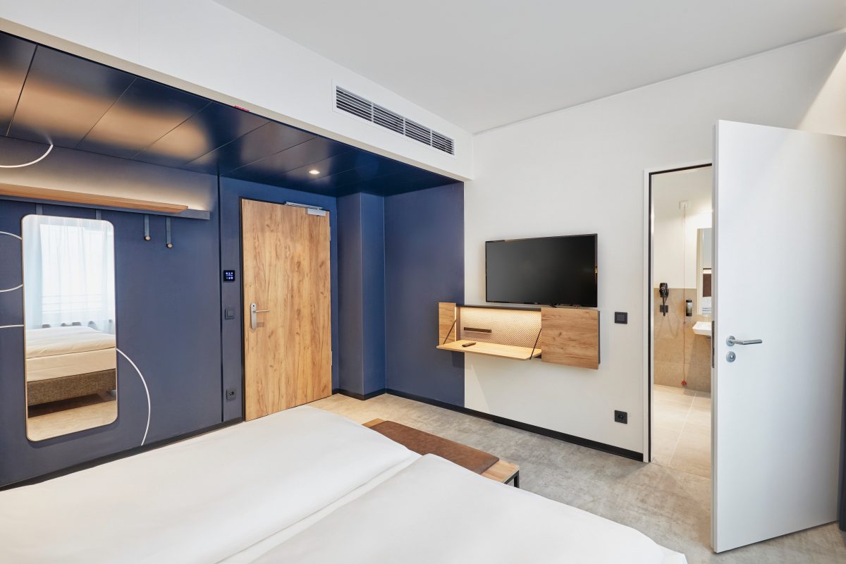 Modernes barrierefreies Hotelzimmer mit Holzdetails, Klapptisch, TV und Zugang zum Bad.
