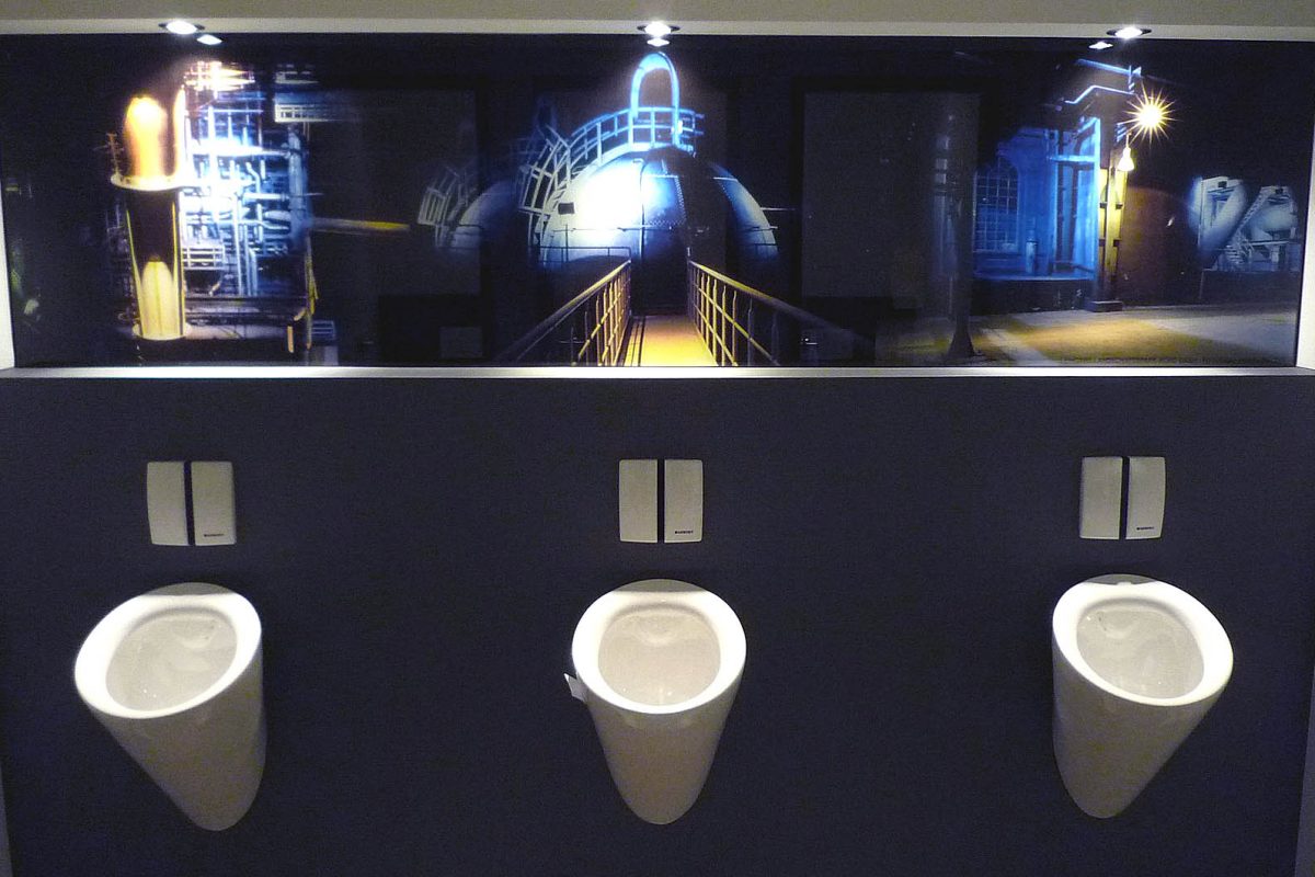 Drei Urinale vor Wandbild mit nächtlicher Industrieanlagen-Szene.