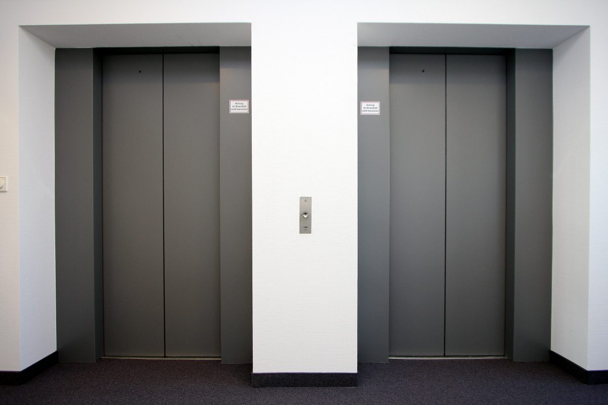 Zwei graue Aufzugtüren in heller Wandnische mit Ruftaste dazwischen.