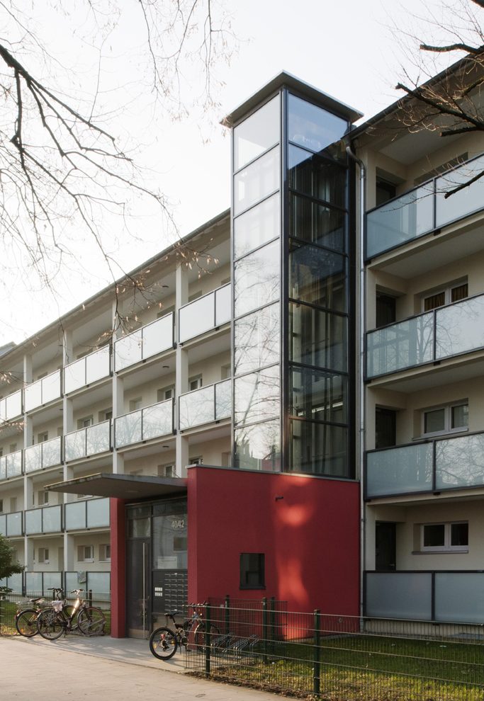 Eingangsbereich eines sanierten Wohnhauses mit Glasaufzug und roten Fassadenelementen.