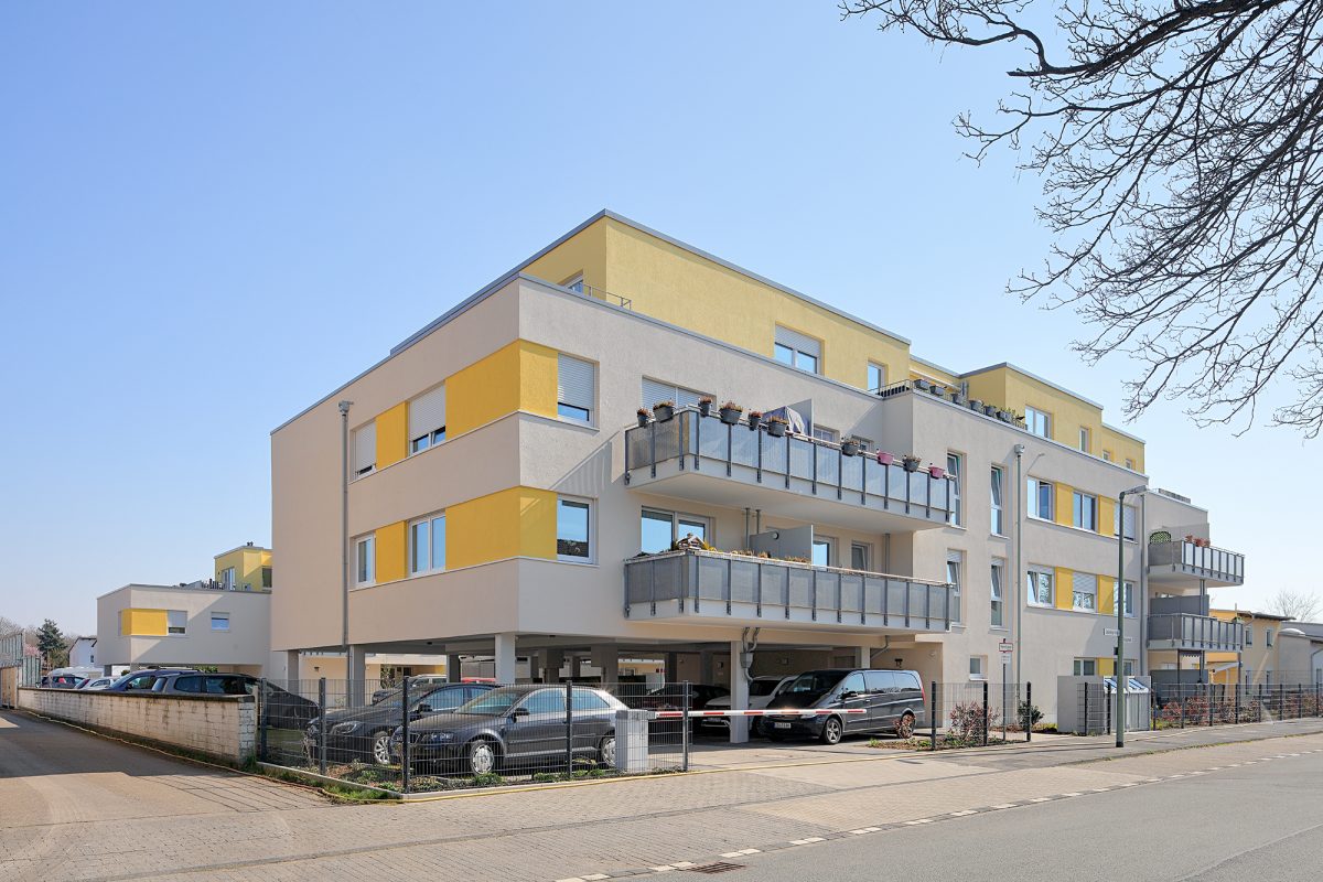 Moderner Wohnbau mit gelben Fassadenteilen und Stellplätzen im Erdgeschoss.