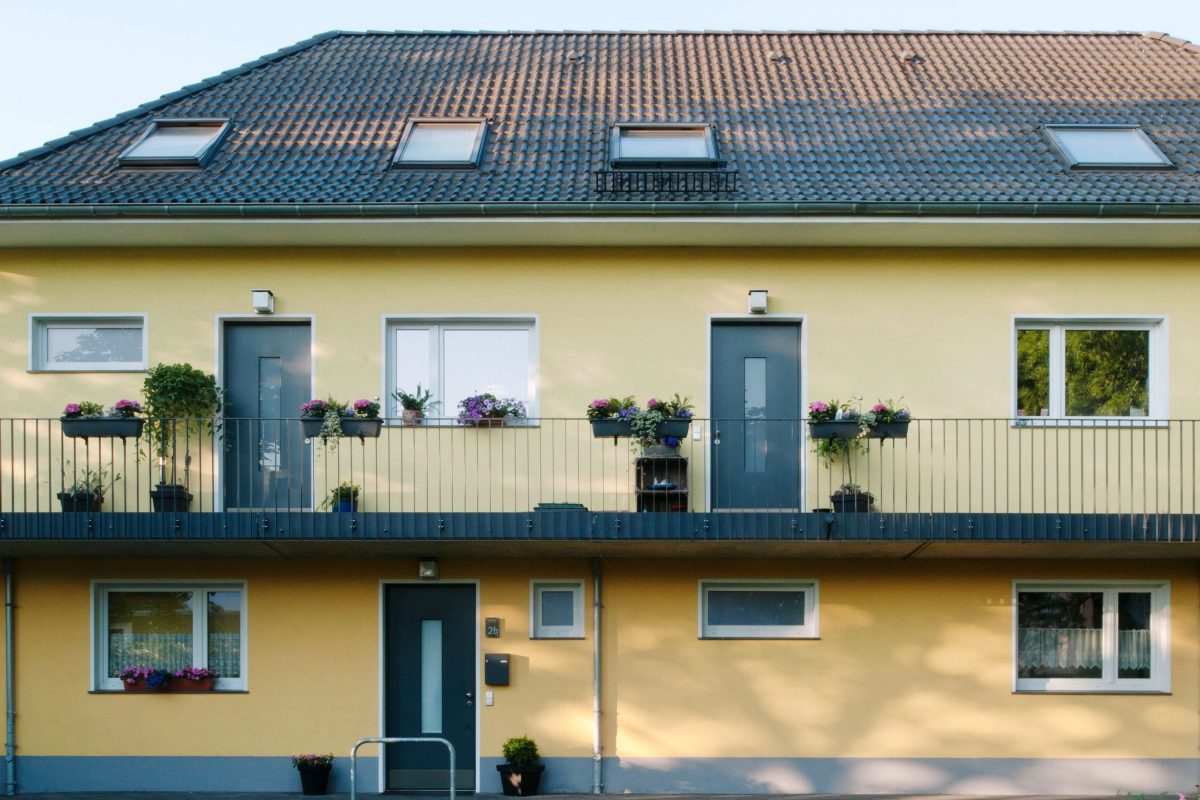 Gelbes Mehrfamilienhaus mit Laubengang und blumigem Geländer.