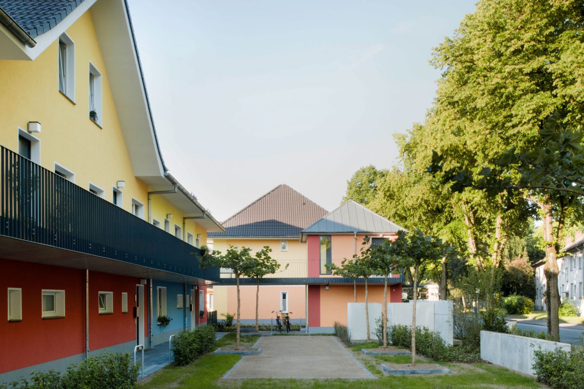 Straßenseitiger Innenhof mit Wegen, Rasen und barrierefreiem Zugang zu bunten Häusern.