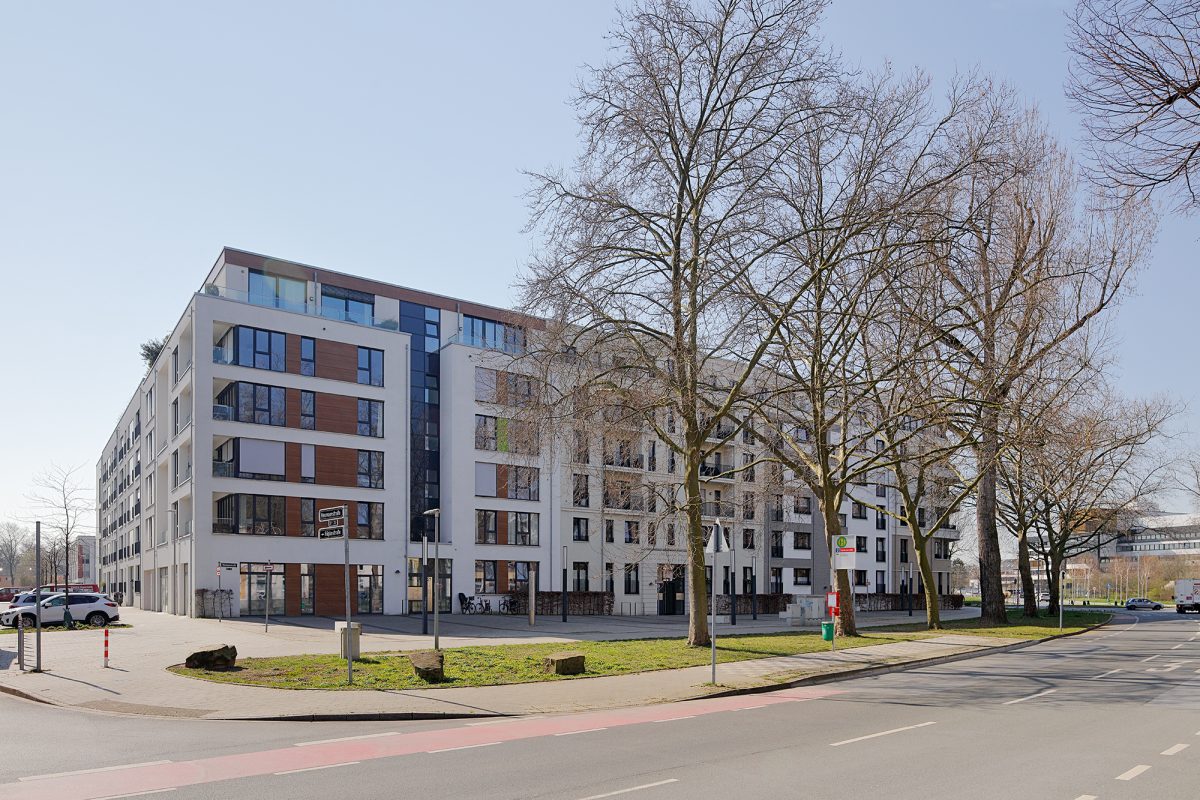 Großes Eckwohnhaus mit Holzfassaden-Elementen an einer Hauptstraße mit großen Bäumen.