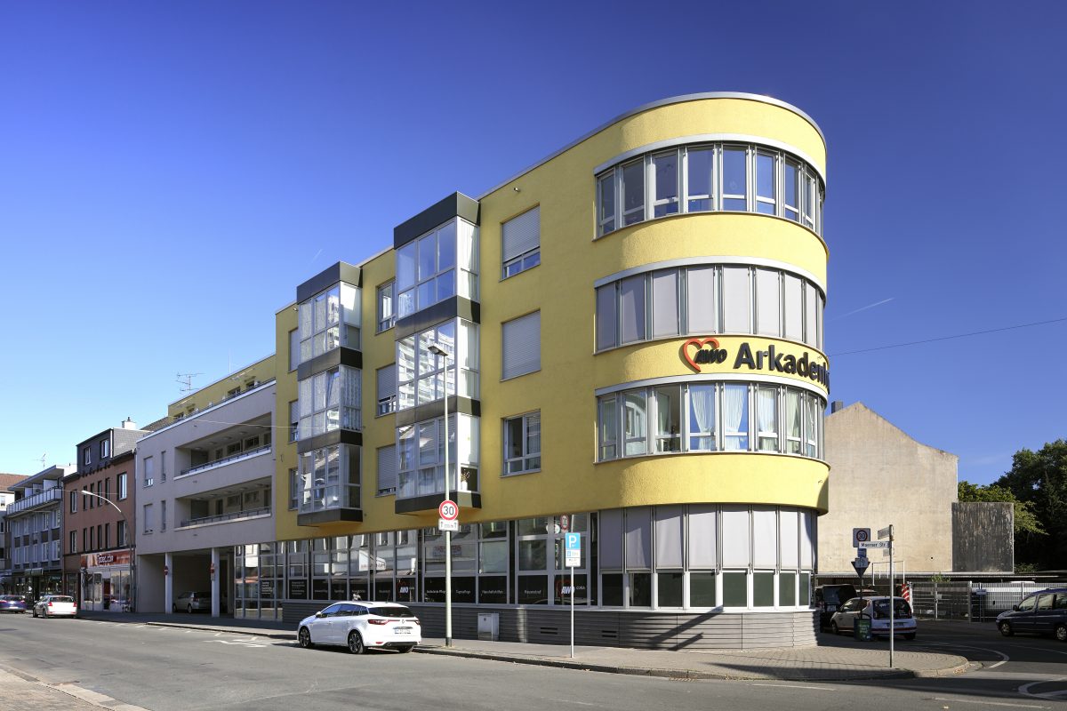 Gelbes Eckgebäude mit Aufschrift „Arkadenhof“ und runder Glasfront.