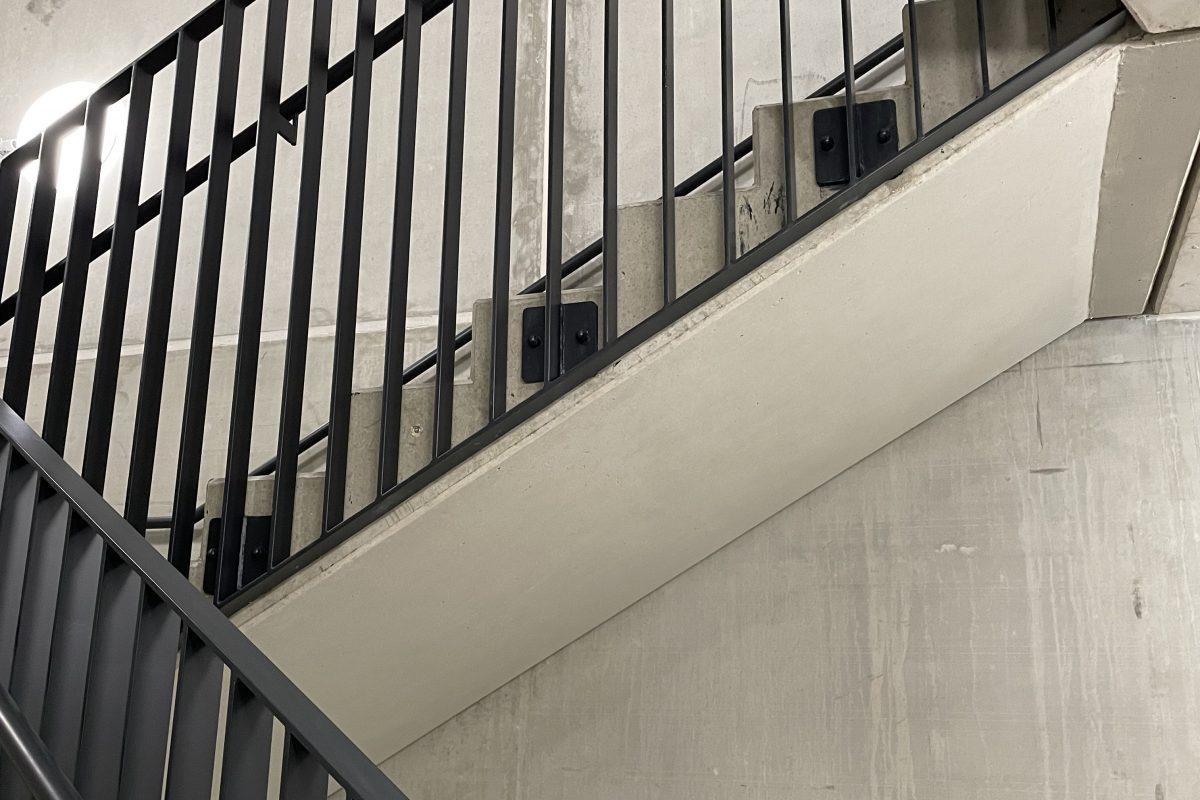 Betontreppe mit schwarzem Metallgeländer vor rauer Sichtbetonwand.