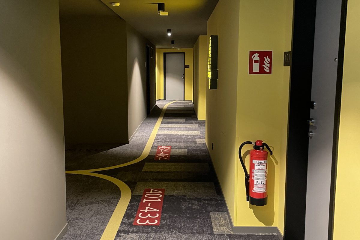 Hotelgang mit gelben Wänden und Teppich mit Wegmarkierungen.