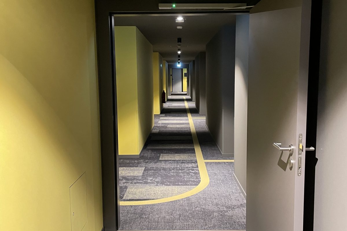 Hotelgang mit offener Tür, gelben Wänden und Teppich mit Wegmarkierungen.