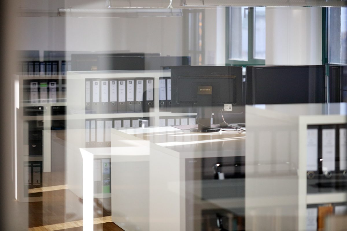 Blick durch Glaswand auf modernes Büro mit Monitoren und Aktenordnern.