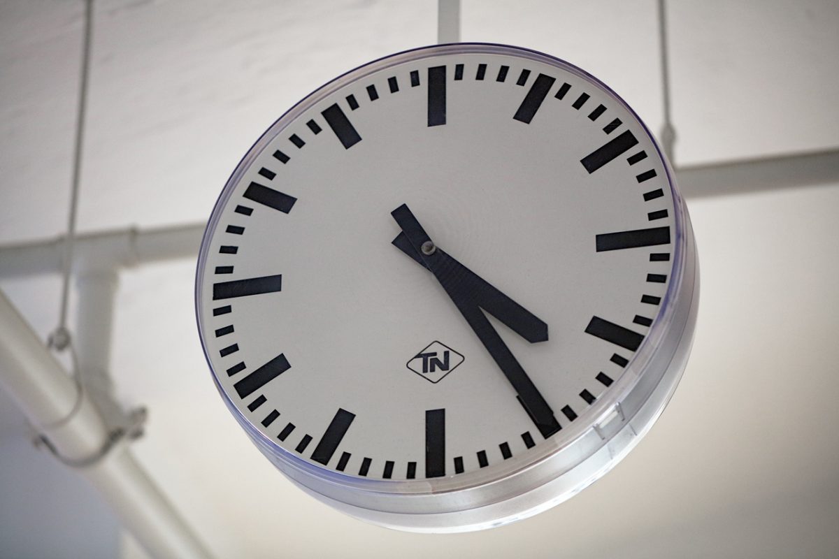 Wanduhr im Bahnhofstil zeigt 16:25 Uhr, schlichtes Design mit TN-Logo.