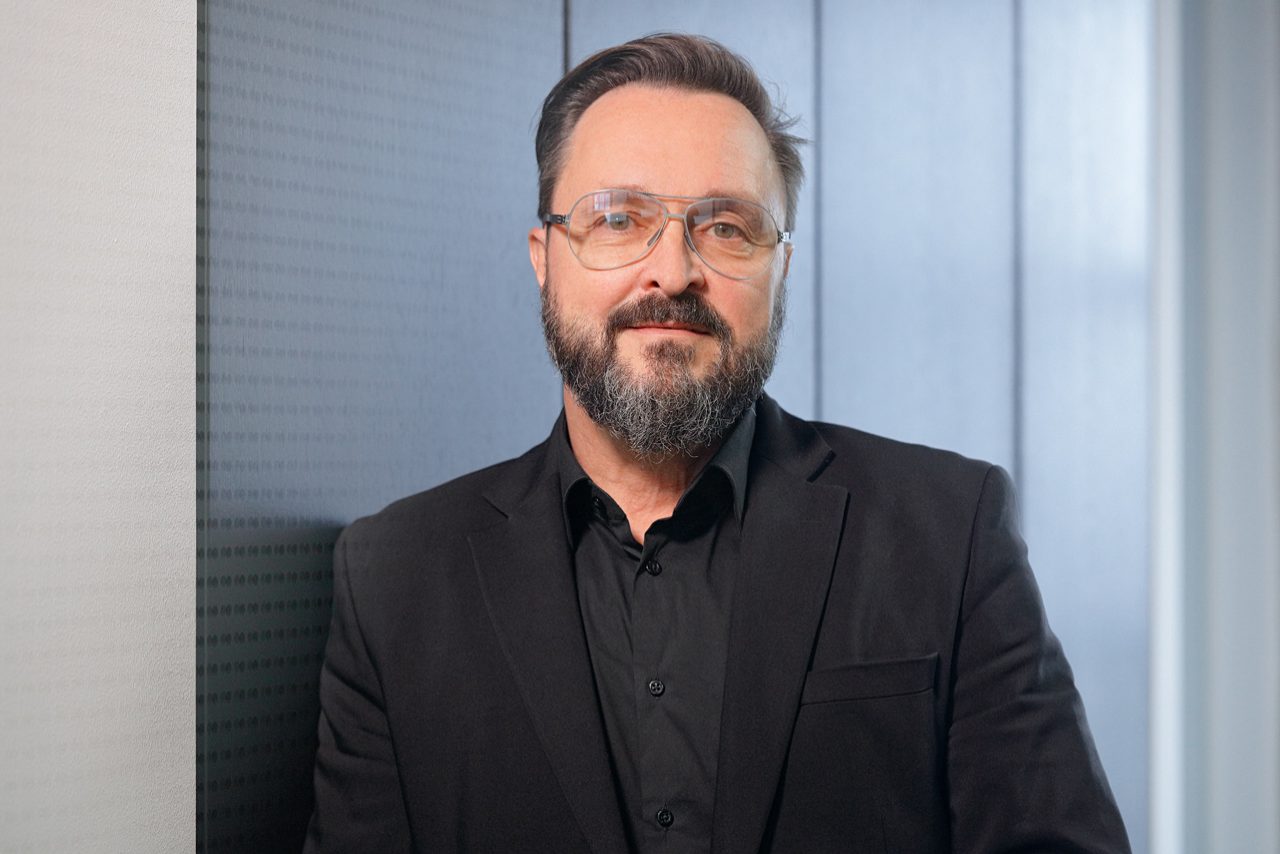 Mann mit Brille und Bart im schwarzen Anzug vor modernem Hintergrund.