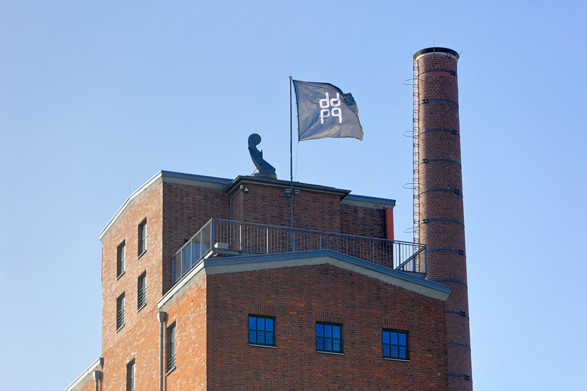 Backsteingebäude mit Schornstein und wehender ddpq-Fahne auf dem Dach