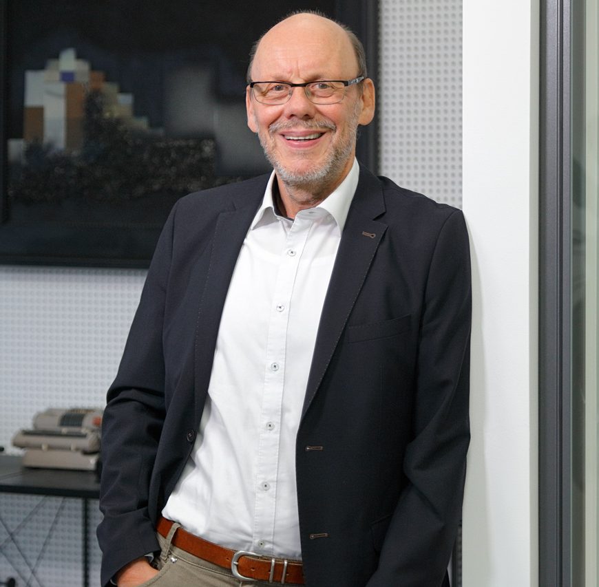 Ein freundlich lächelnder Mann im Anzug steht in modernem Büroambiente.