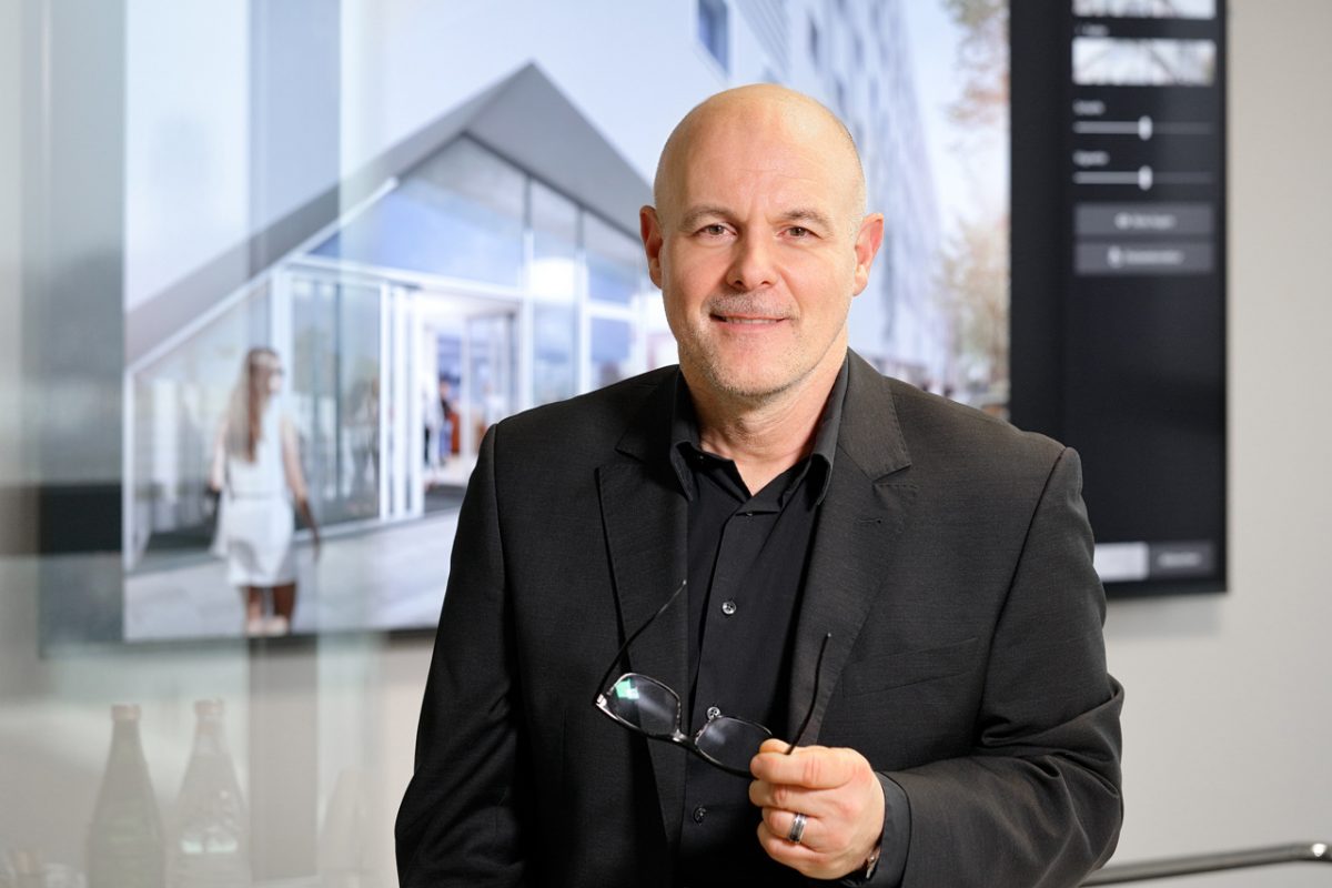 Mann im dunklen Anzug mit Brille in der Hand vor Architekturbildschirm