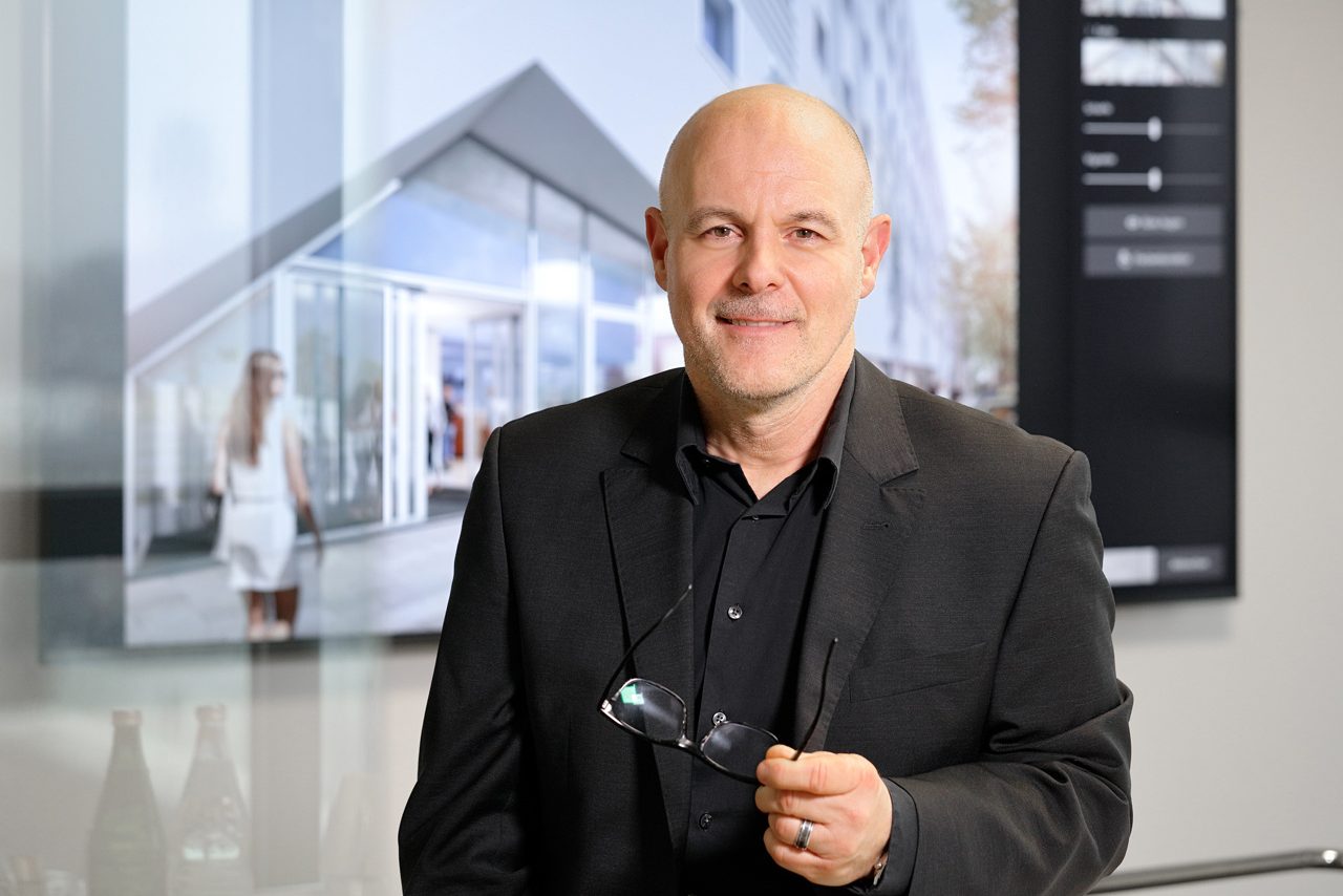 Mann im dunklen Anzug mit Brille in der Hand vor Architekturbildschirm