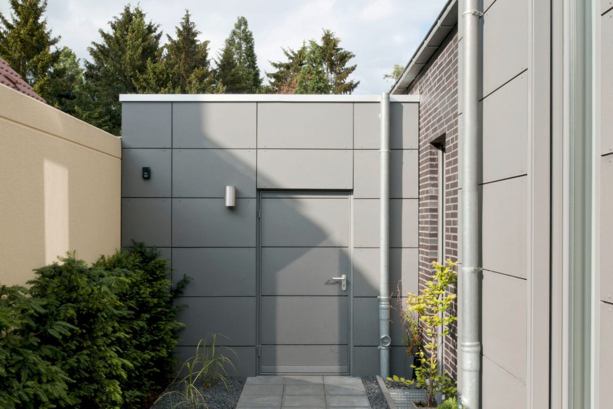 Seitlicher Hauszugang mit grauer Vorhangfassade und Pflasterweg.